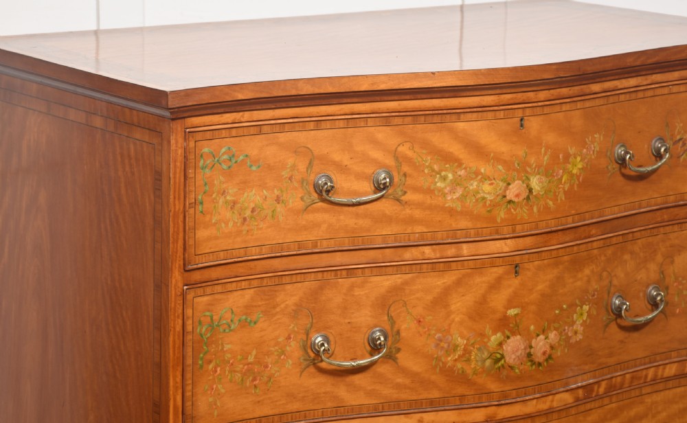 A Sheraton Revival satinwood chest of drawers featuring a serpentine front, three drawers decorated with painted floral swags, and brass handles. View III