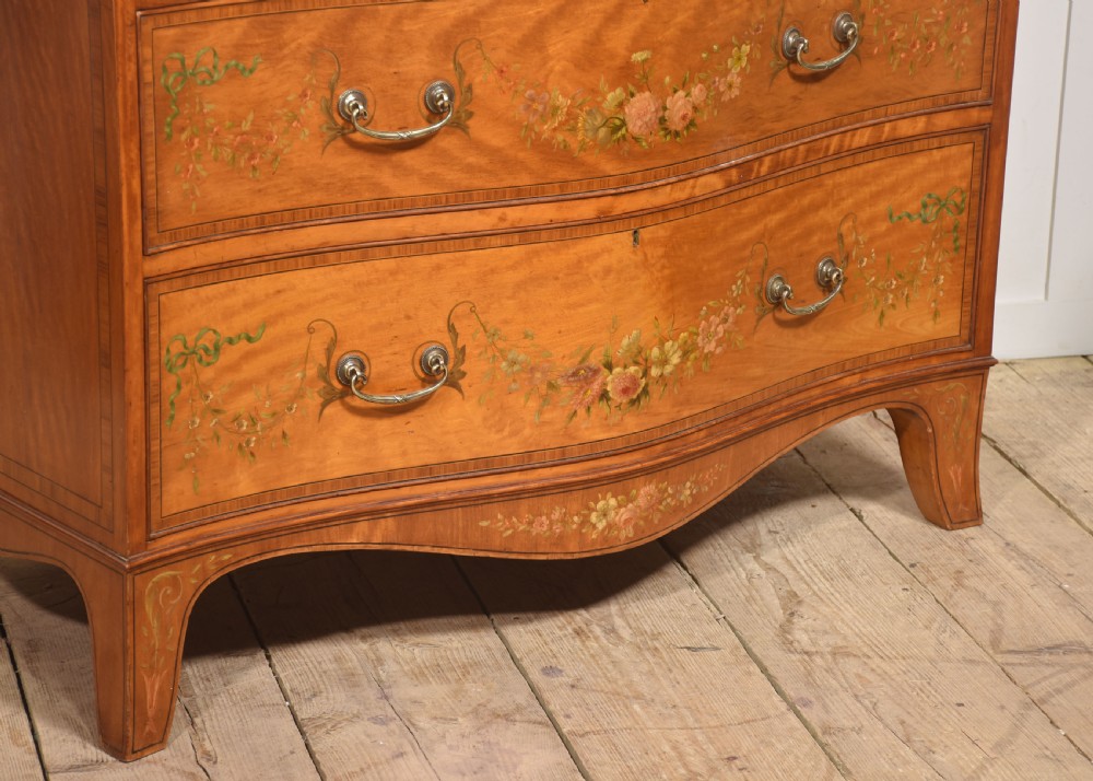 A Sheraton Revival satinwood chest of drawers featuring a serpentine front, three drawers decorated with painted floral swags, and brass handles. View V