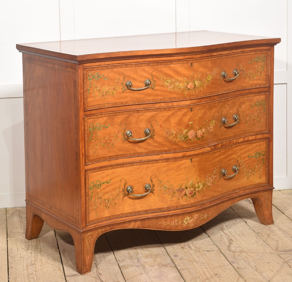 A Sheraton Revival satinwood chest of drawers featuring a serpentine front, three drawers decorated with painted floral swags, and brass handles. View VI