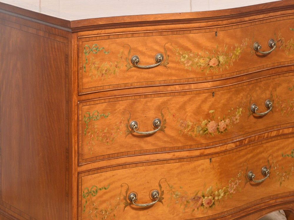 A Sheraton Revival satinwood chest of drawers featuring a serpentine front, three drawers decorated with painted floral swags, and brass handles. View VII