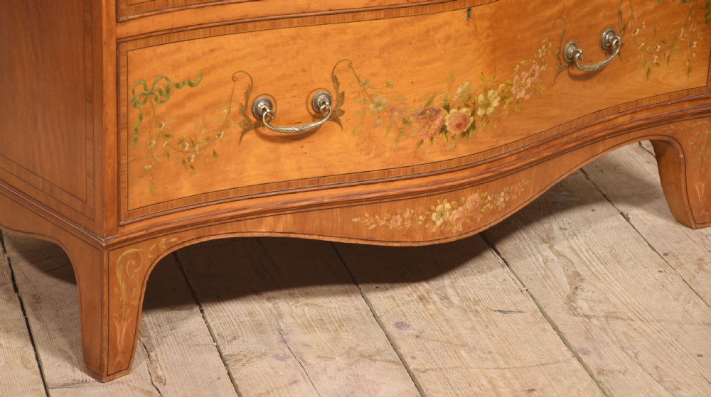 A Sheraton Revival satinwood chest of drawers featuring a serpentine front, three drawers decorated with painted floral swags, and brass handles. View VIII