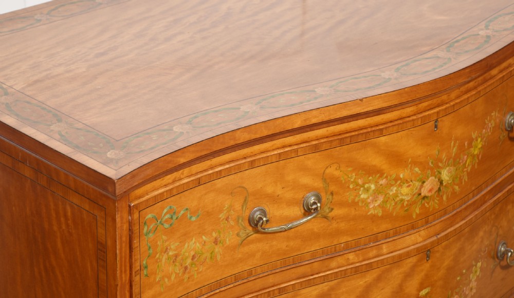 A Sheraton Revival satinwood chest of drawers featuring a serpentine front, three drawers decorated with painted floral swags, and brass handles. View IX