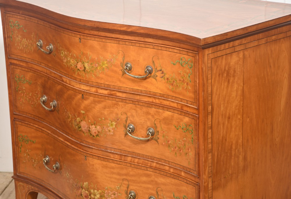 A Sheraton Revival satinwood chest of drawers featuring a serpentine front, three drawers decorated with painted floral swags, and brass handles. View XIII