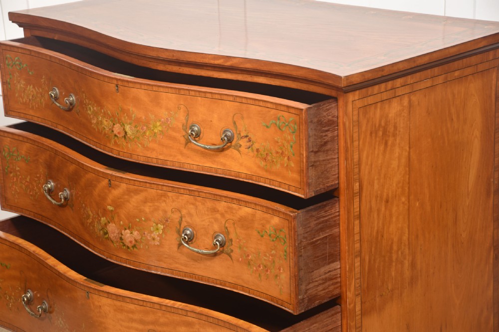 A Sheraton Revival satinwood chest of drawers featuring a serpentine front, three drawers decorated with painted floral swags, and brass handles. View XIV