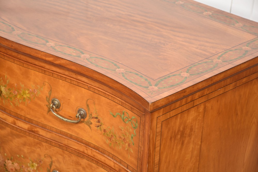 A Sheraton Revival satinwood chest of drawers featuring a serpentine front, three drawers decorated with painted floral swags, and brass handles. View XV