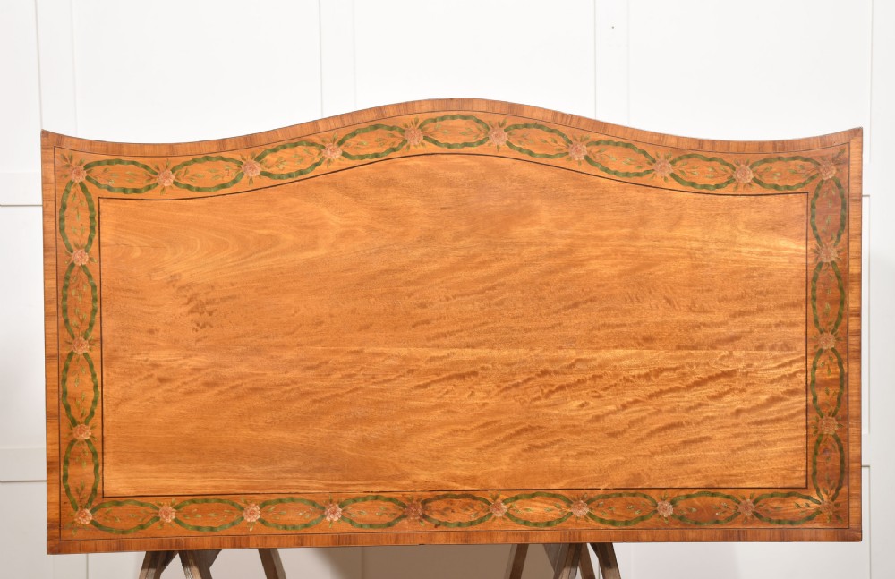 A Sheraton Revival satinwood chest of drawers featuring a serpentine front, three drawers decorated with painted floral swags, and brass handles. View XVI