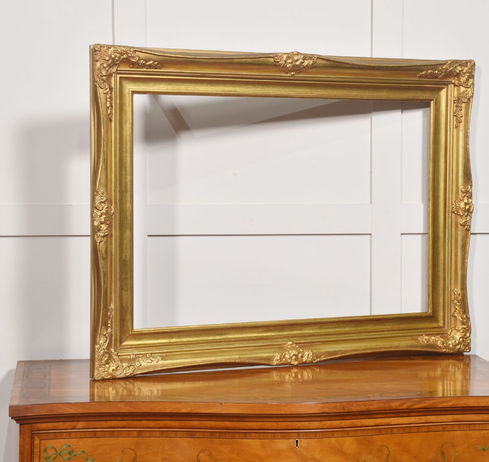 An ornate rectangular giltwood picture frame in the Rococo style featuring decorative corner mouldings and swept edges.
