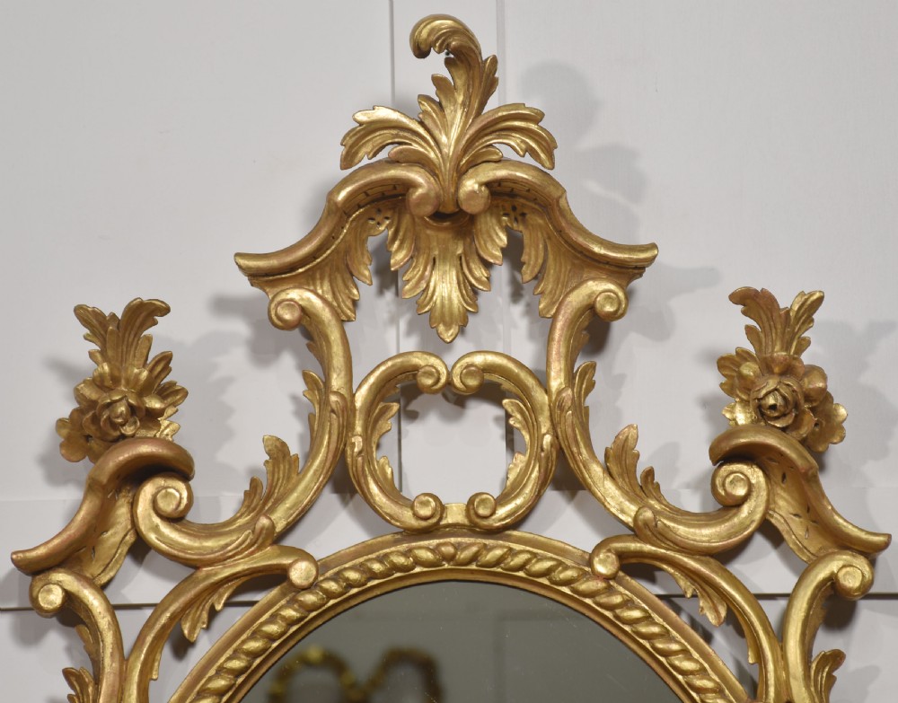 An oval wall mirror set in a gilded frame decorated with carved C-scrolls, floral motifs, and an acanthus leaf crest. View II