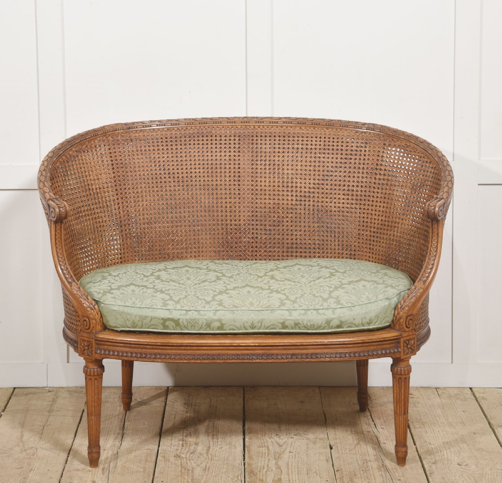 A Louis XVI Revival carved walnut settee featuring a curved woven cane back and a green damask upholstered seat cushion, supported by turned fluted legs.