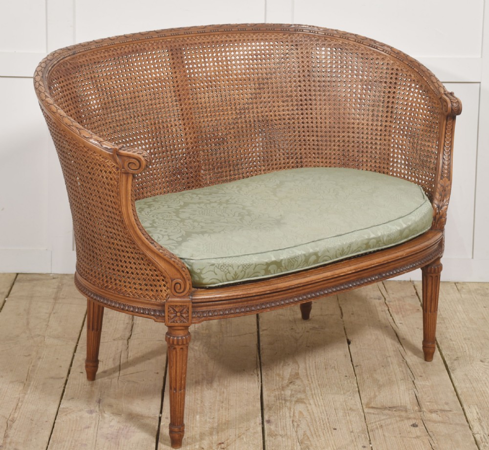 A Louis XVI Revival carved walnut settee featuring a curved woven cane back and a green damask upholstered seat cushion, supported by turned fluted legs. View IV