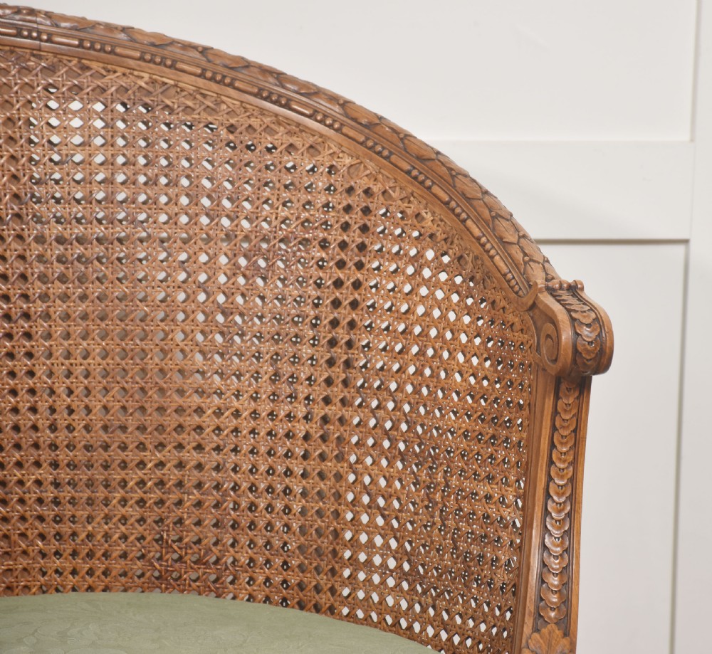A Louis XVI Revival carved walnut settee featuring a curved woven cane back and a green damask upholstered seat cushion, supported by turned fluted legs. View VI