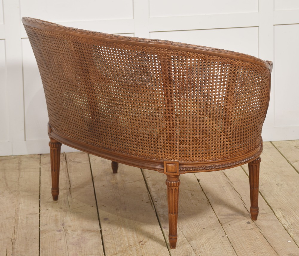 A Louis XVI Revival carved walnut settee featuring a curved woven cane back and a green damask upholstered seat cushion, supported by turned fluted legs. View IX
