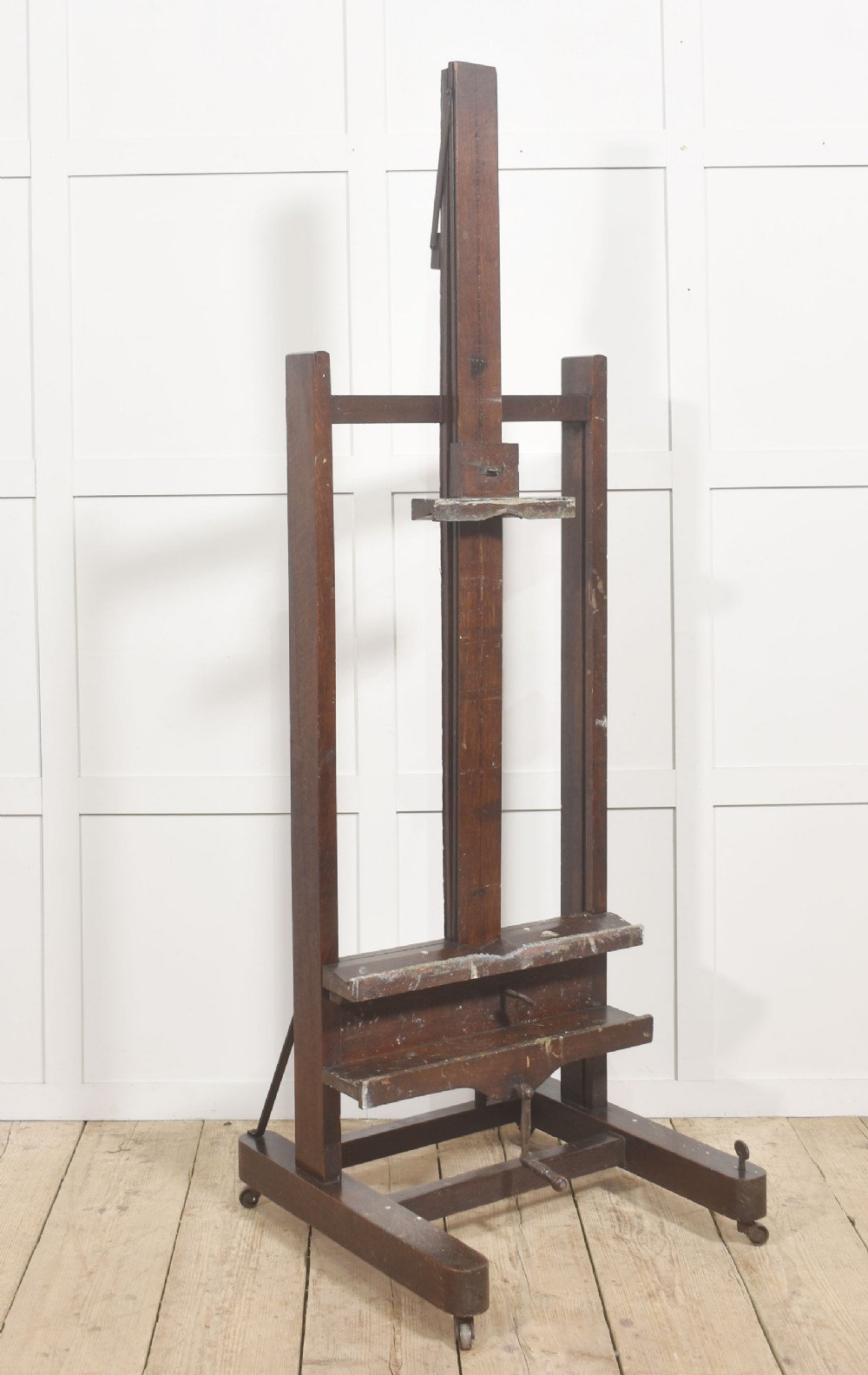 A tall dark oak adjustable artists studio easel featuring a hand-crank winding mechanism and castors.