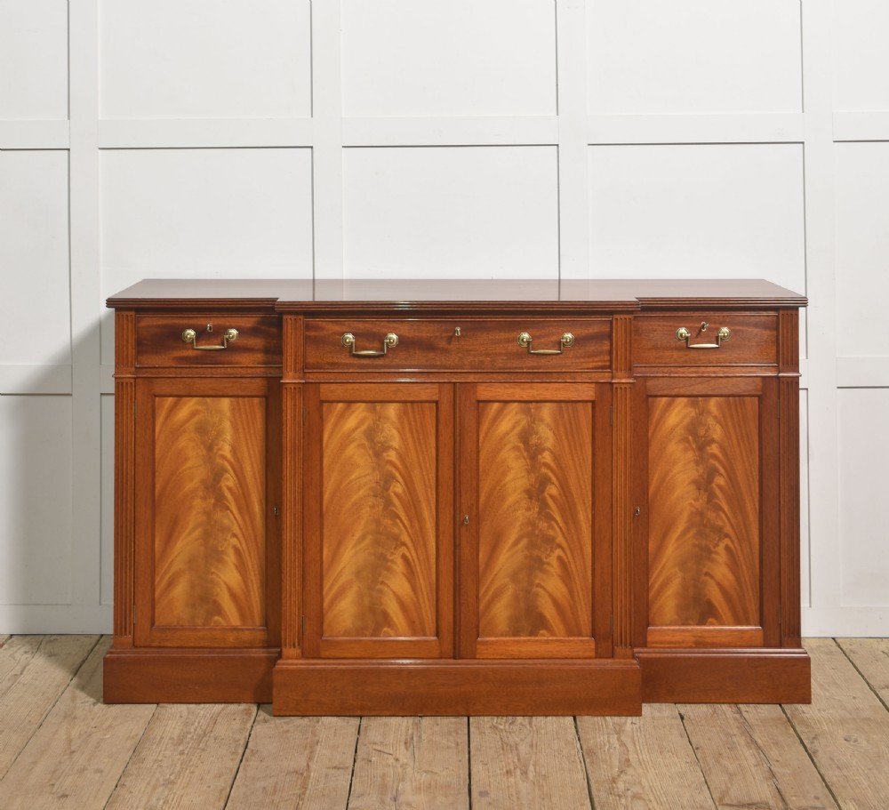 A flame mahogany breakfront sideboard featuring four panelled cupboard doors, four top drawers with brass drop handles, and fluted pilasters.