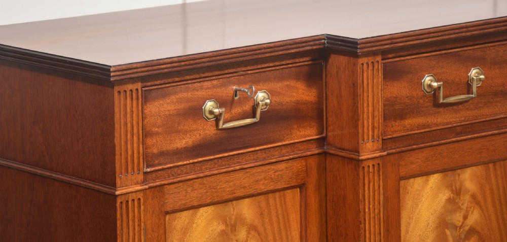 A flame mahogany breakfront sideboard featuring four panelled cupboard doors, four top drawers with brass drop handles, and fluted pilasters. View IV