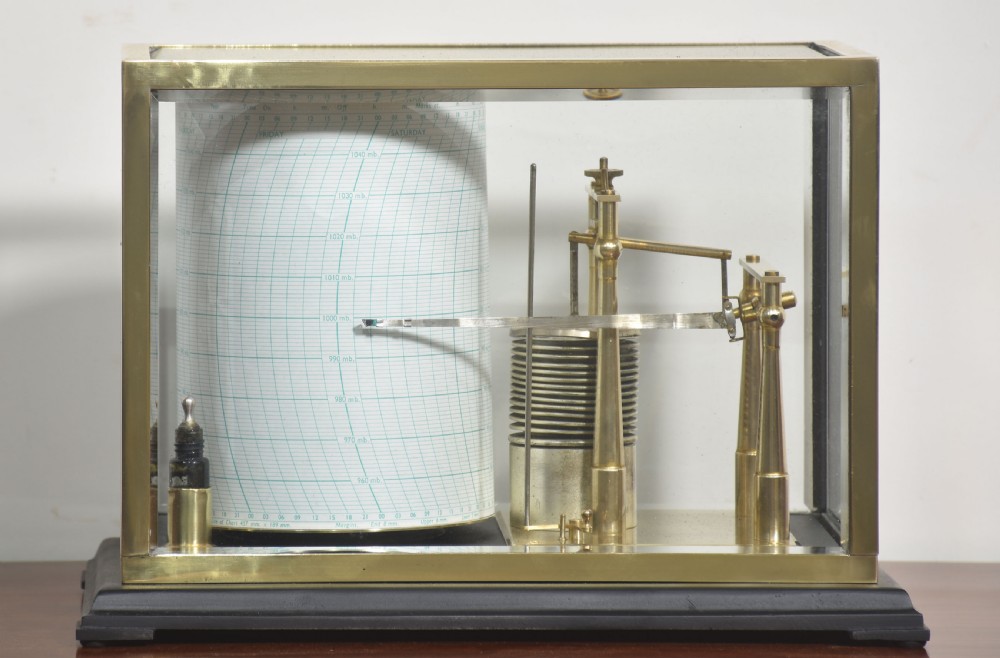 An antique brass-framed glass display case housing a scientific barograph mechanism with a paper chart drum, aneroid capsules, and pen arm.