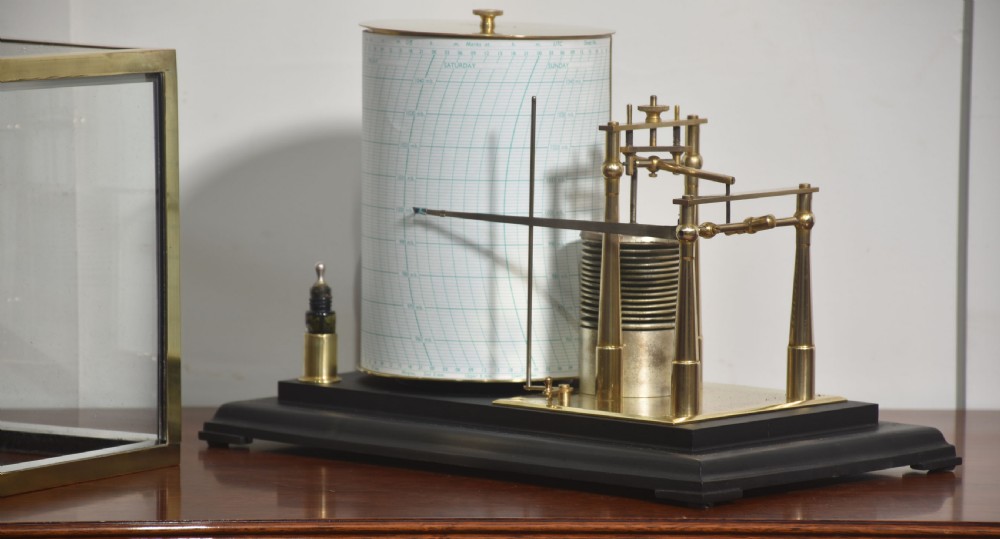 An antique brass-framed glass display case housing a scientific barograph mechanism with a paper chart drum, aneroid capsules, and pen arm. View VI