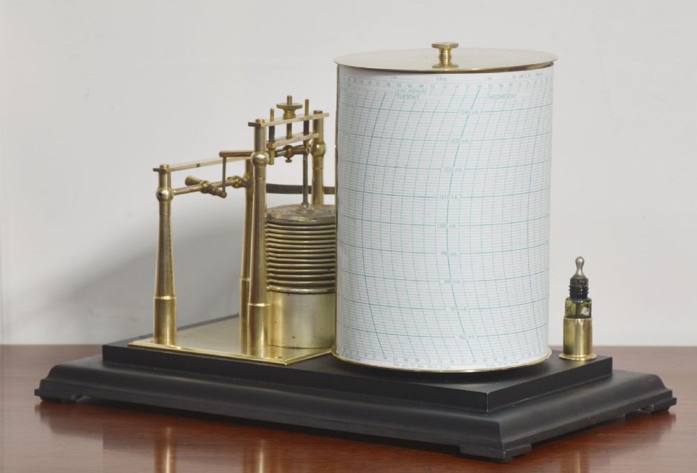 An antique brass-framed glass display case housing a scientific barograph mechanism with a paper chart drum, aneroid capsules, and pen arm. View X