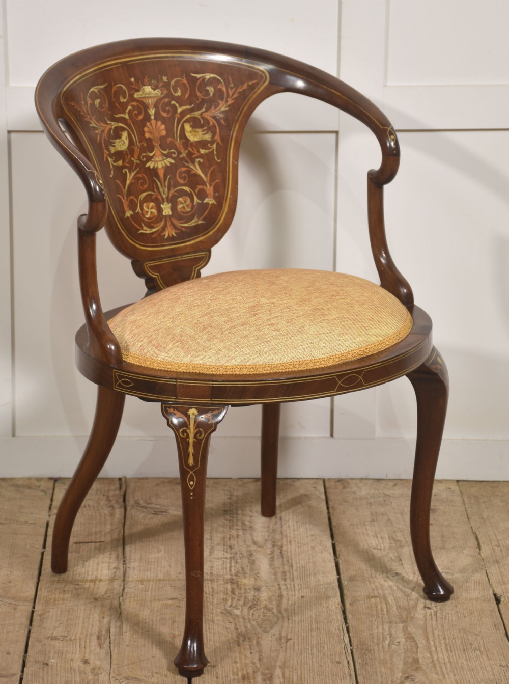 An Edwardian mahogany tub armchair displaying foliate marquetry inlay across the curved back splat and cabriole legs, paired with a textured cream seat.