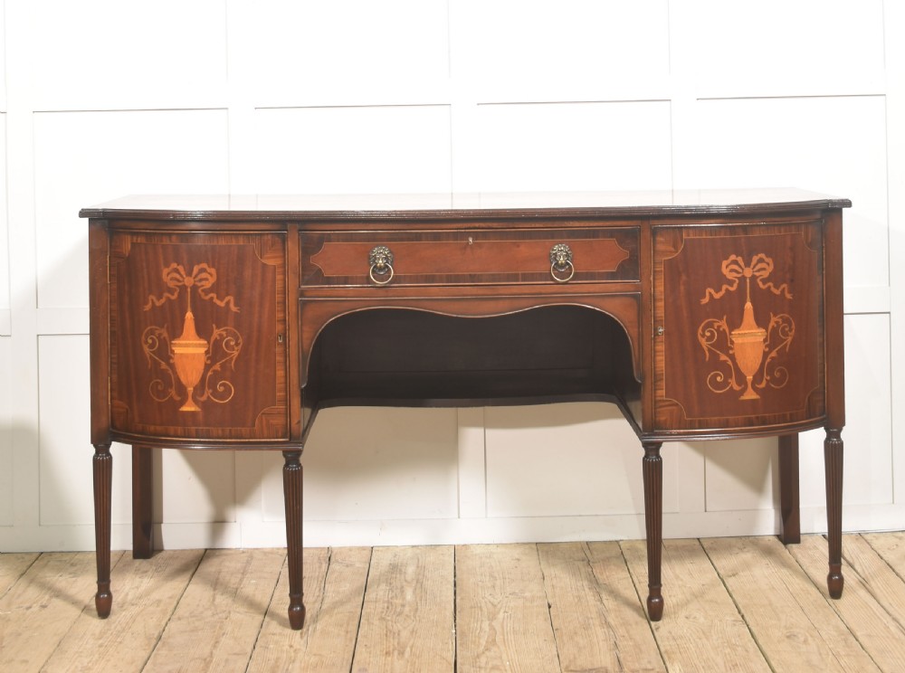 A George III style mahogany bow-front sideboard featuring marquetry urn inlays on twin side cupboards, a central drawer with brass lion mask handles, and reeded tapering legs.