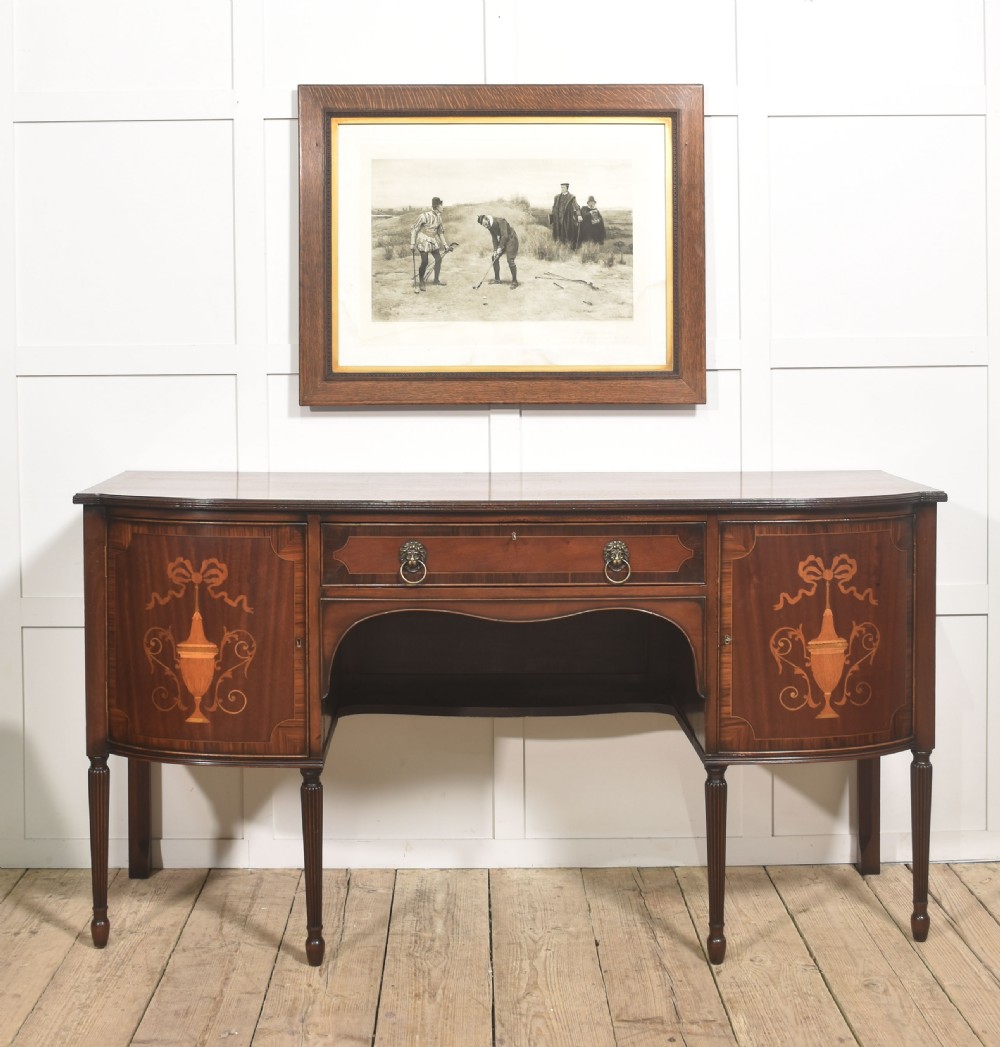 A George III style mahogany bow-front sideboard featuring marquetry urn inlays on twin side cupboards, a central drawer with brass lion mask handles, and reeded tapering legs. View II