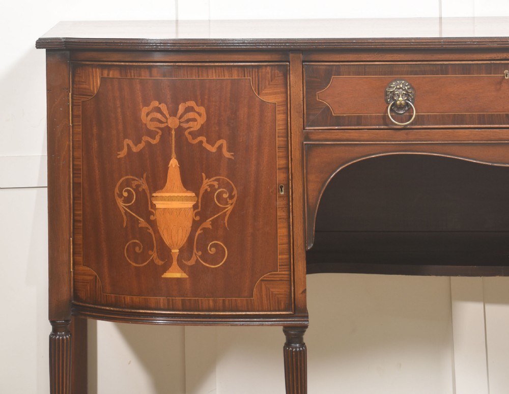 A George III style mahogany bow-front sideboard featuring marquetry urn inlays on twin side cupboards, a central drawer with brass lion mask handles, and reeded tapering legs. View III