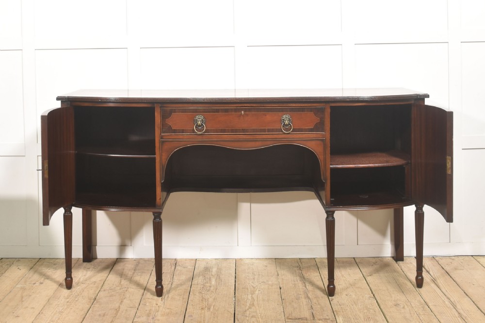 A George III style mahogany bow-front sideboard featuring marquetry urn inlays on twin side cupboards, a central drawer with brass lion mask handles, and reeded tapering legs. View V