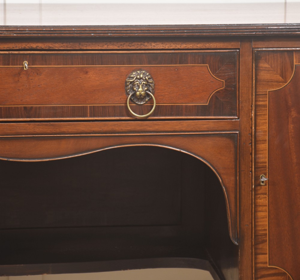 A George III style mahogany bow-front sideboard featuring marquetry urn inlays on twin side cupboards, a central drawer with brass lion mask handles, and reeded tapering legs. View VI