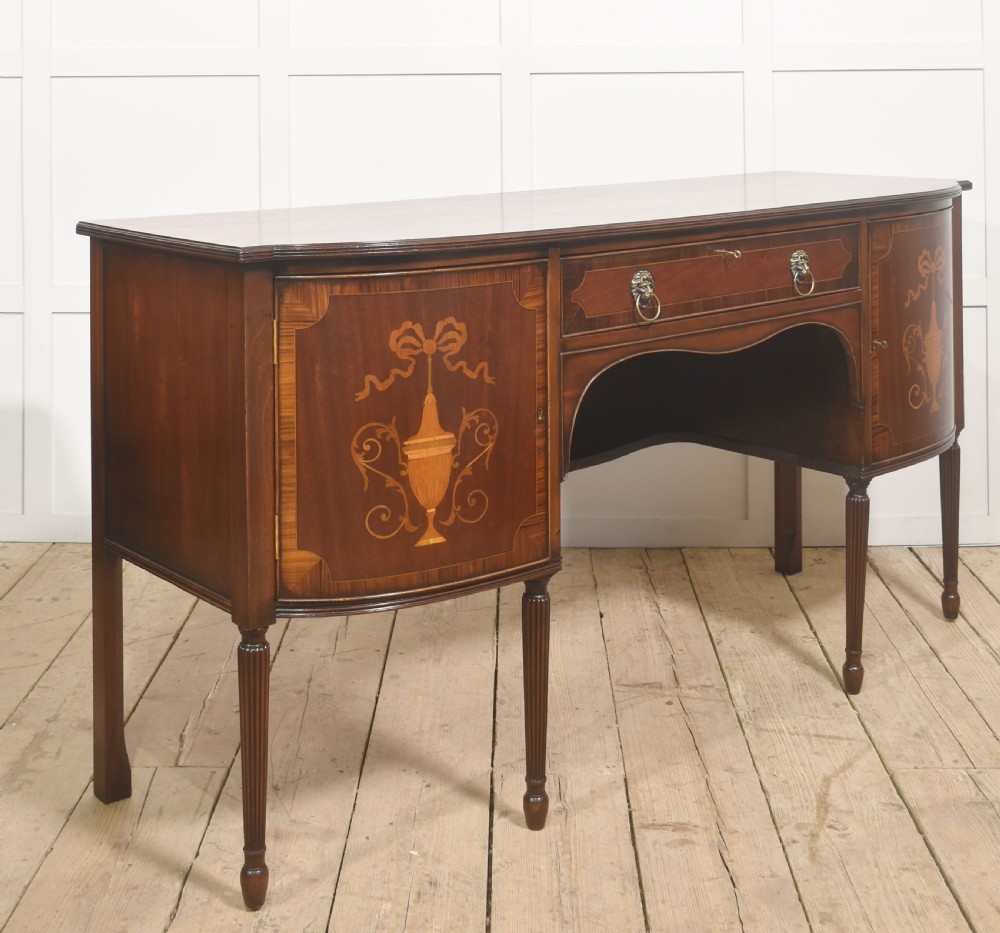 A George III style mahogany bow-front sideboard featuring marquetry urn inlays on twin side cupboards, a central drawer with brass lion mask handles, and reeded tapering legs. View VII