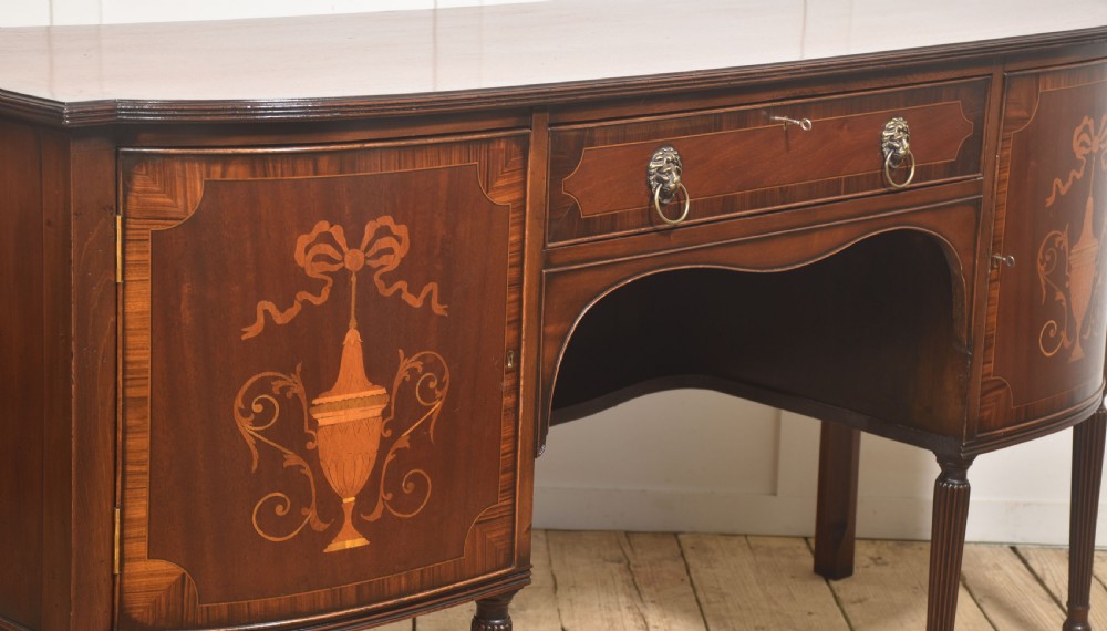 A George III style mahogany bow-front sideboard featuring marquetry urn inlays on twin side cupboards, a central drawer with brass lion mask handles, and reeded tapering legs. View VIII