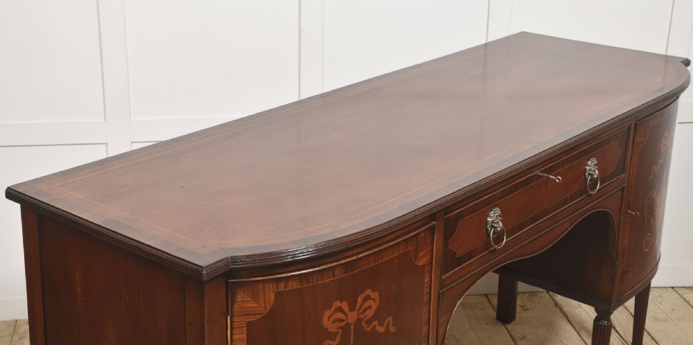 A George III style mahogany bow-front sideboard featuring marquetry urn inlays on twin side cupboards, a central drawer with brass lion mask handles, and reeded tapering legs. View XI