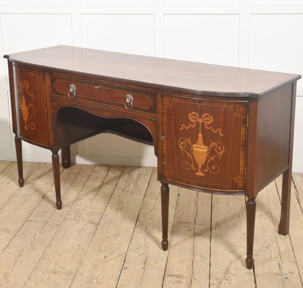 A George III style mahogany bow-front sideboard featuring marquetry urn inlays on twin side cupboards, a central drawer with brass lion mask handles, and reeded tapering legs. View XII