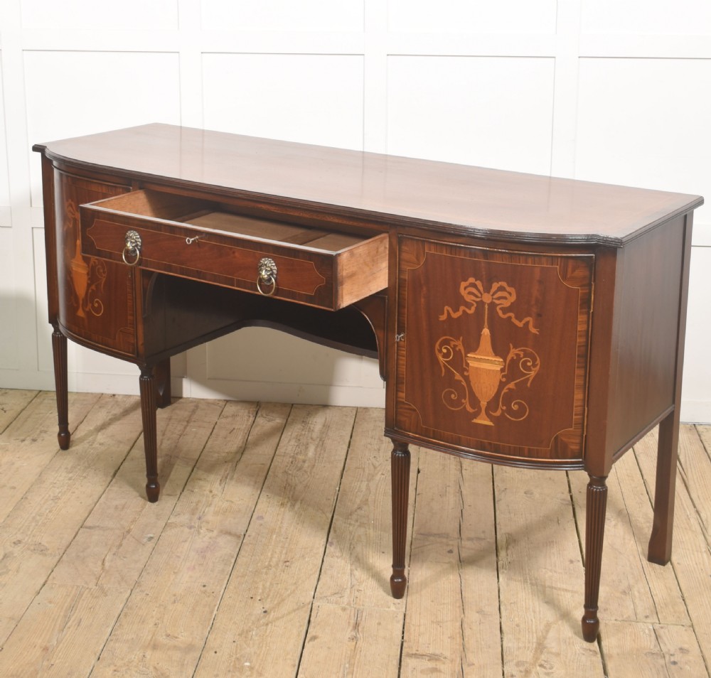 A George III style mahogany bow-front sideboard featuring marquetry urn inlays on twin side cupboards, a central drawer with brass lion mask handles, and reeded tapering legs. View XIII
