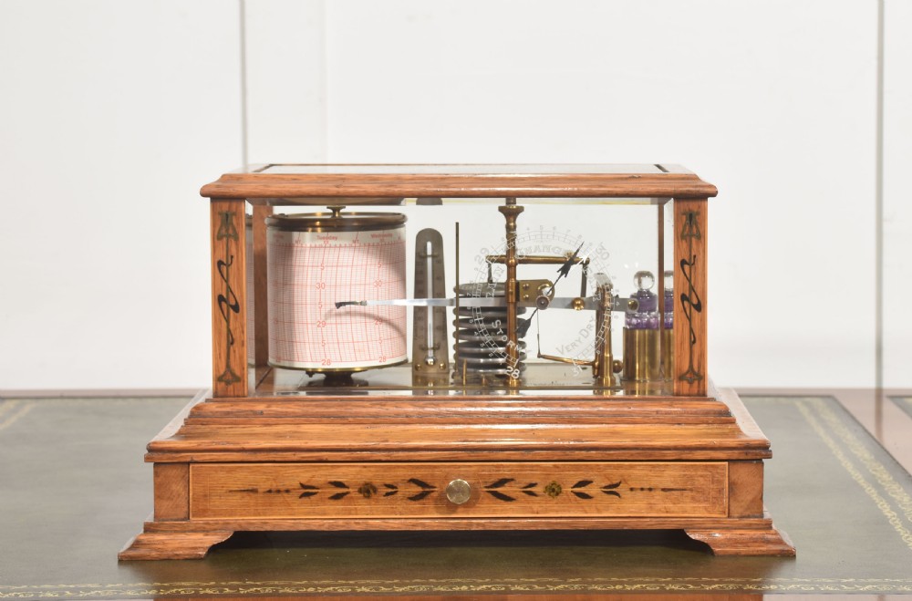 An Edwardian oak-cased barograph featuring bevelled glass panels, polished brass movement, paper chart drum, and floral Art Nouveau wood inlay along the base drawer and corner pillars.