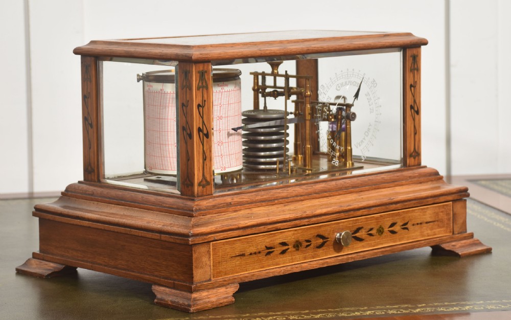 An Edwardian oak-cased barograph featuring bevelled glass panels, polished brass movement, paper chart drum, and floral Art Nouveau wood inlay along the base drawer and corner pillars. View III