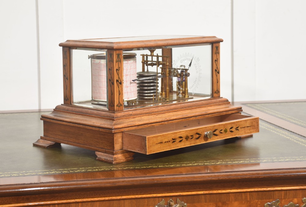 An Edwardian oak-cased barograph featuring bevelled glass panels, polished brass movement, paper chart drum, and floral Art Nouveau wood inlay along the base drawer and corner pillars. View VII