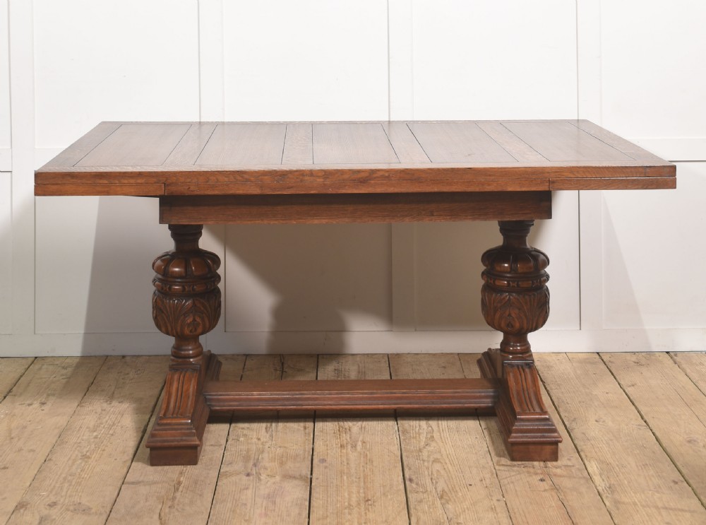 A solid oak refectory dining table featuring a rectangular planked top supported by two carved cup-and-cover pedestal legs connected by a central stretcher.