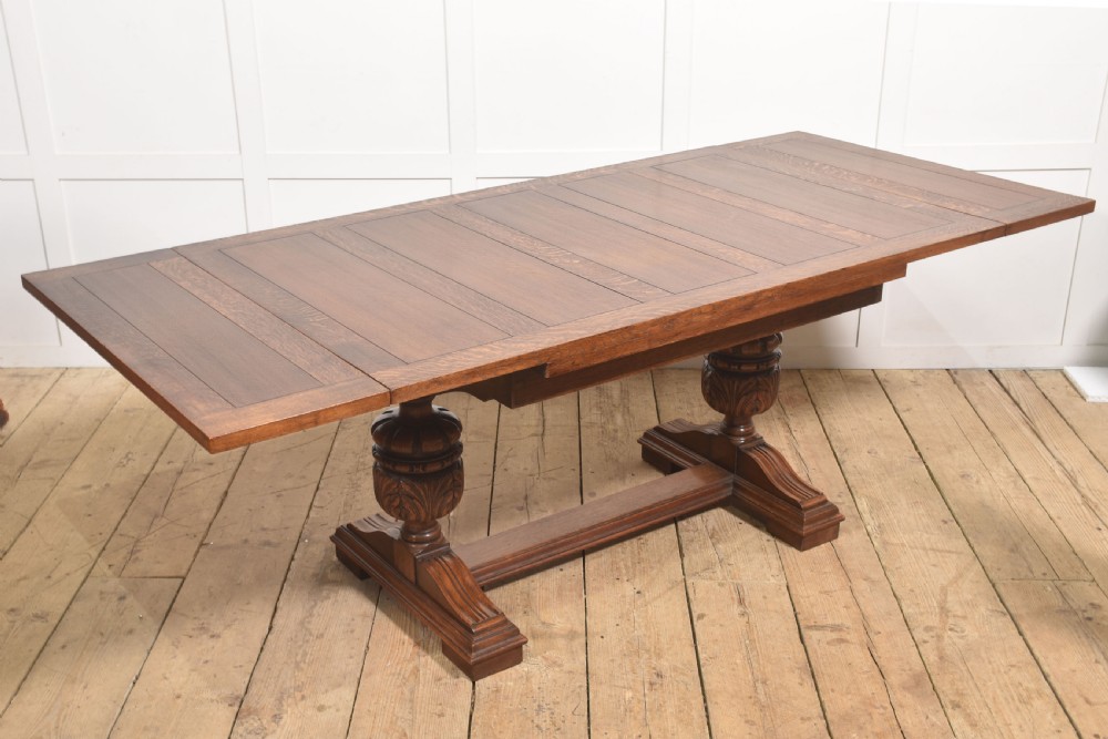 A solid oak refectory dining table featuring a rectangular planked top supported by two carved cup-and-cover pedestal legs connected by a central stretcher. View VI