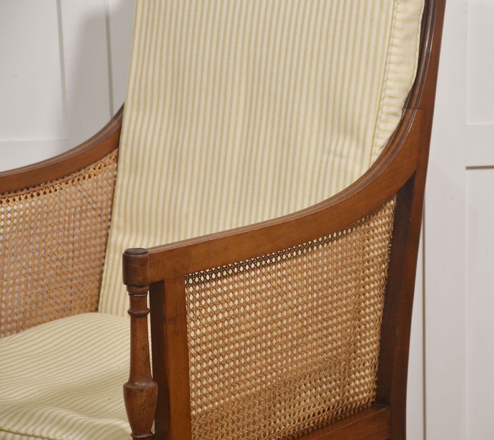 A Regency mahogany bergere armchair featuring woven cane back and side panels, turned front legs on brass castors, and light striped fabric cushions. View II