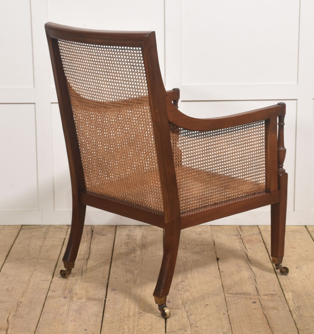 A Regency mahogany bergere armchair featuring woven cane back and side panels, turned front legs on brass castors, and light striped fabric cushions. View X