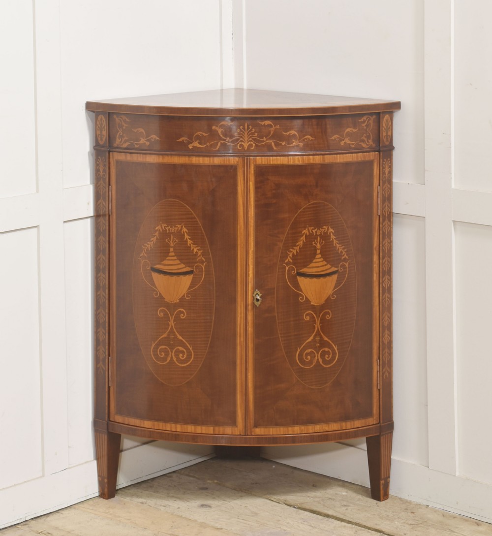 A floor-standing Sheraton Revival bow-front mahogany corner cabinet with satinwood neoclassical urn marquetry on twin doors.