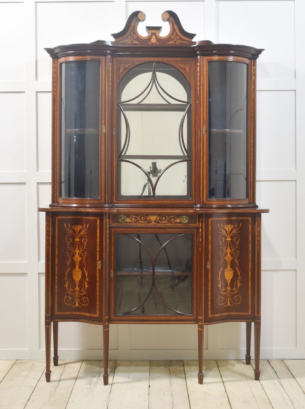 An Edwardian mahogany display cabinet featuring satinwood marquetry inlay, urn motifs, a swan-neck pediment, curved glass doors, and tapered legs.