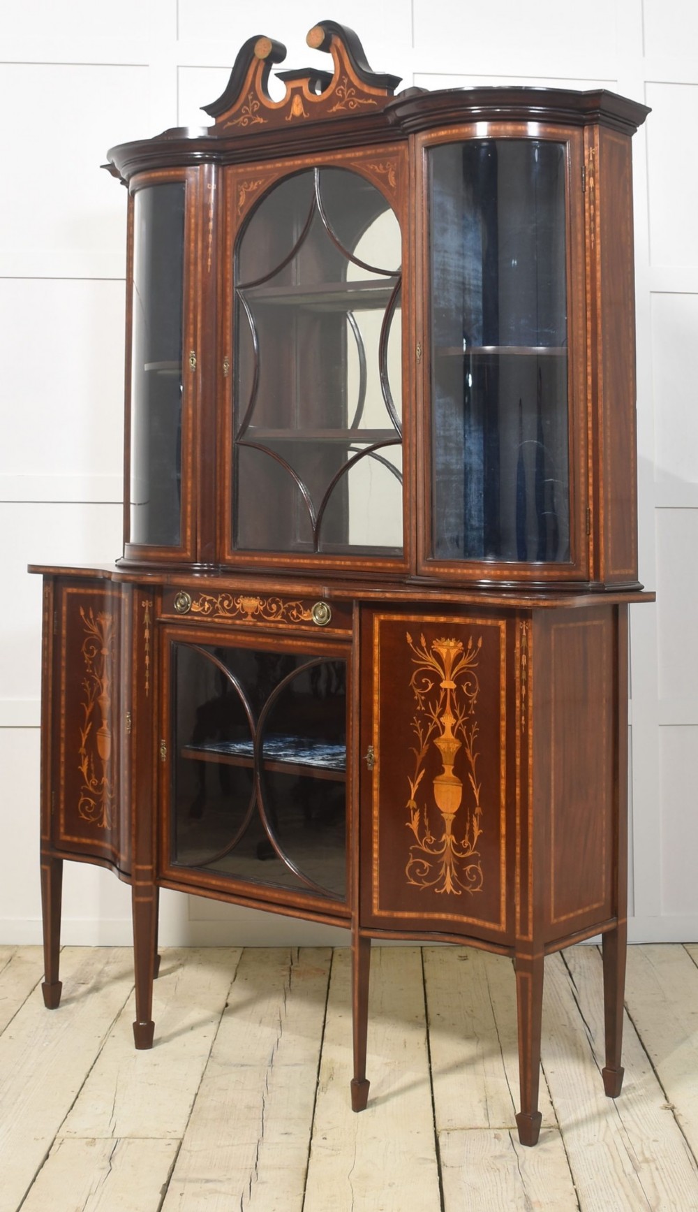 An Edwardian mahogany display cabinet featuring satinwood marquetry inlay, urn motifs, a swan-neck pediment, curved glass doors, and tapered legs. View II