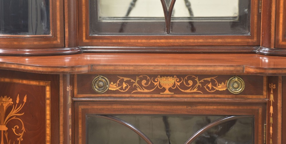 An Edwardian mahogany display cabinet featuring satinwood marquetry inlay, urn motifs, a swan-neck pediment, curved glass doors, and tapered legs. View III