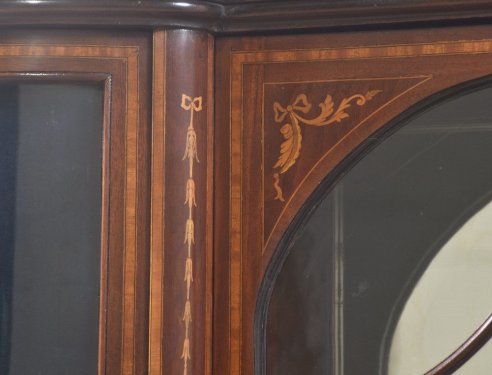 An Edwardian mahogany display cabinet featuring satinwood marquetry inlay, urn motifs, a swan-neck pediment, curved glass doors, and tapered legs. View IV