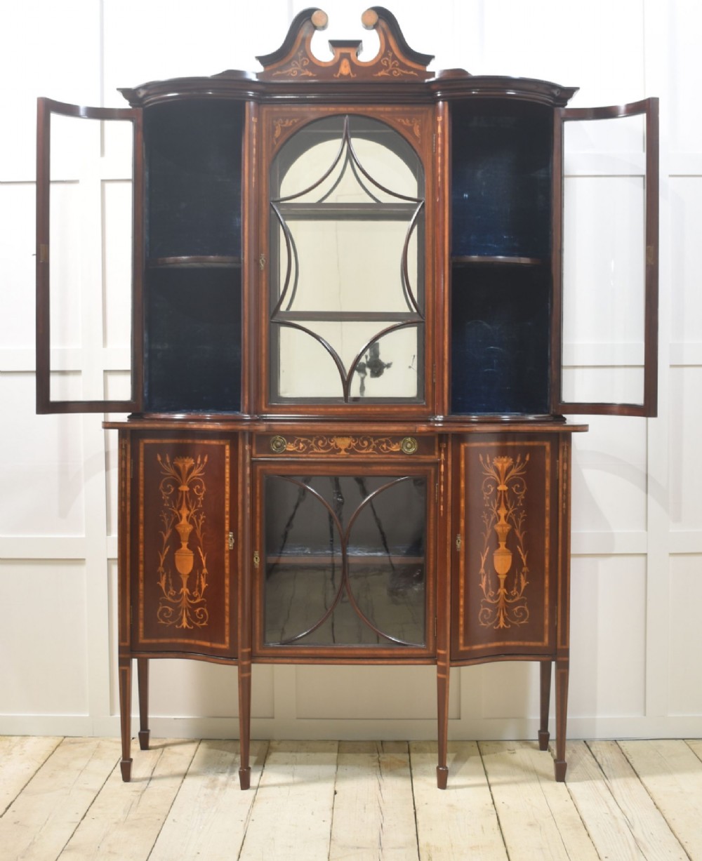 An Edwardian mahogany display cabinet featuring satinwood marquetry inlay, urn motifs, a swan-neck pediment, curved glass doors, and tapered legs. View V