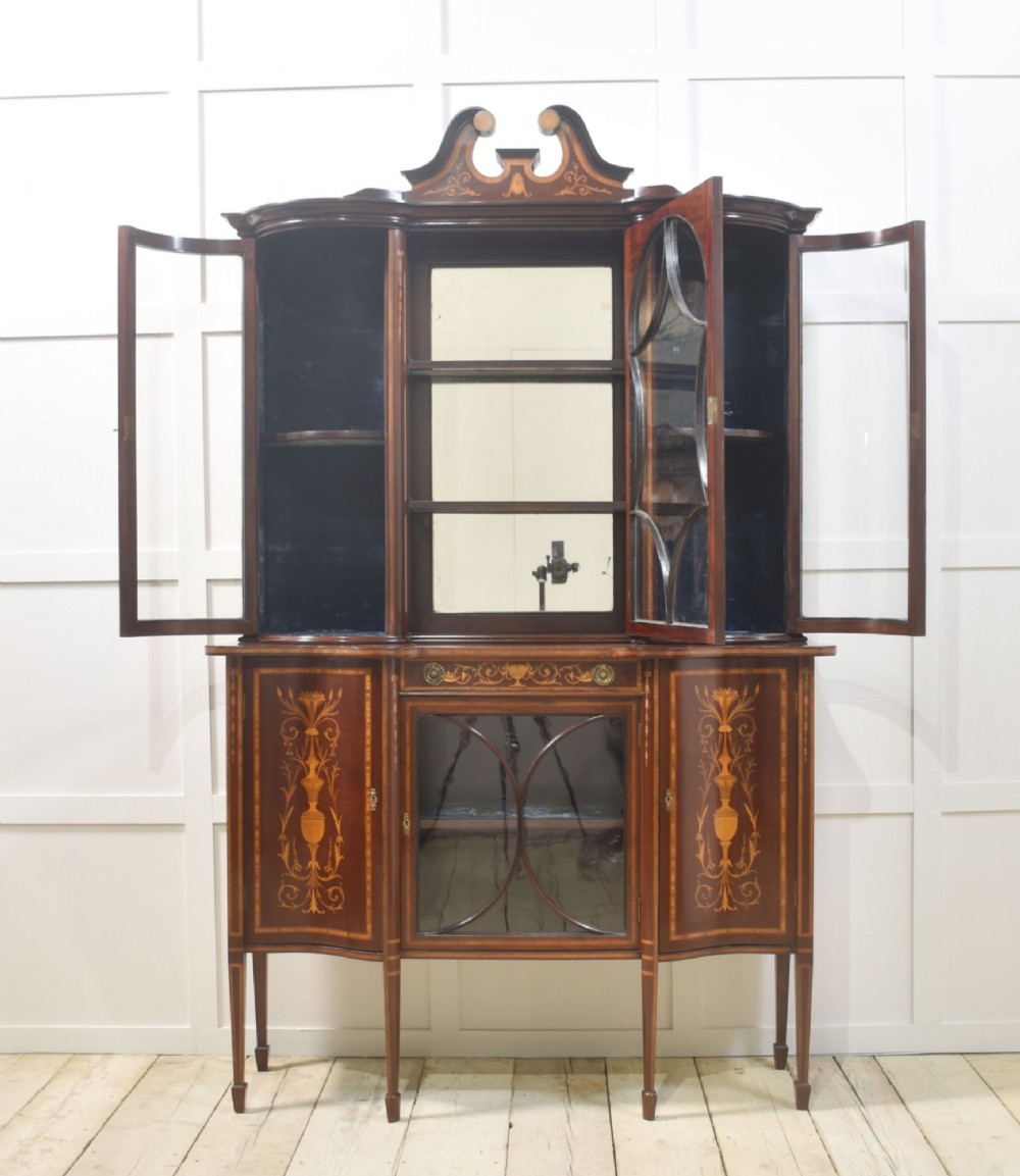 An Edwardian mahogany display cabinet featuring satinwood marquetry inlay, urn motifs, a swan-neck pediment, curved glass doors, and tapered legs. View VI