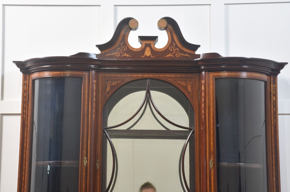 An Edwardian mahogany display cabinet featuring satinwood marquetry inlay, urn motifs, a swan-neck pediment, curved glass doors, and tapered legs. View IX