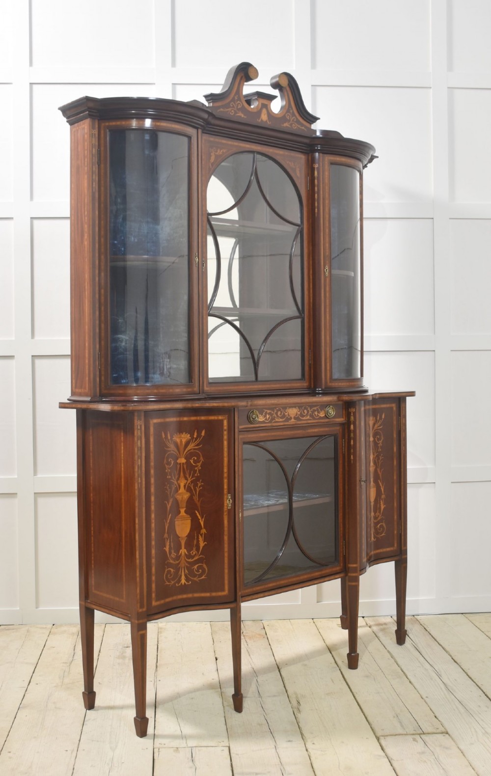 An Edwardian mahogany display cabinet featuring satinwood marquetry inlay, urn motifs, a swan-neck pediment, curved glass doors, and tapered legs. View X