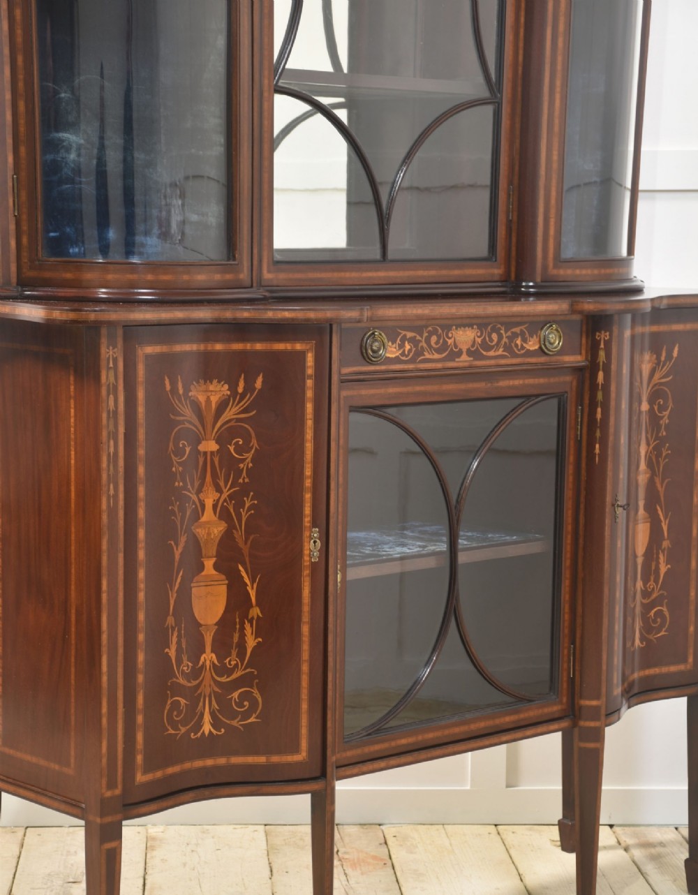 An Edwardian mahogany display cabinet featuring satinwood marquetry inlay, urn motifs, a swan-neck pediment, curved glass doors, and tapered legs. View XI